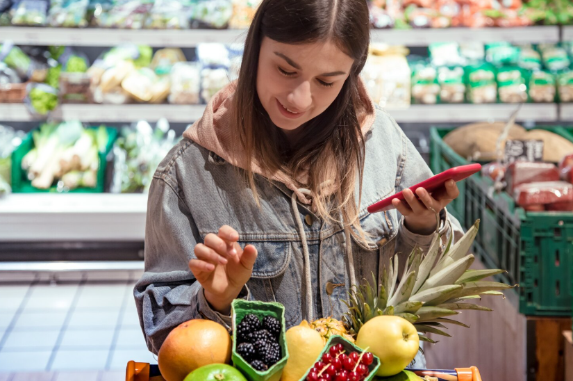 Woman checking grocery prices on her phone while shopping midweek to take advantage of pricing patterns.